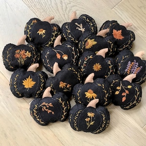 May include: A collection of small, black fabric pumpkins featuring intricate embroidery of various autumn-themed botanical designs, including leaves, flowers, and sprigs, each topped with a small, light-colored fabric stem.