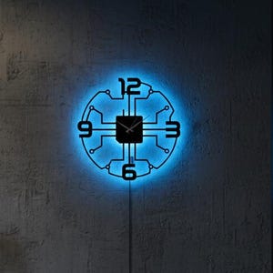 May include: A black, circular wall clock with a circuit board design, illuminated by a blue LED light. The clock features the numbers 12, 3, 6, and 9. The clock is mounted on a grey concrete wall.