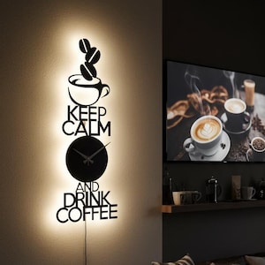 May include: Black wall clock with the words "Keep Calm and Drink Coffee" in a decorative font. The clock features a coffee cup, coffee beans, and a round clock face. The clock is illuminated by a warm light.