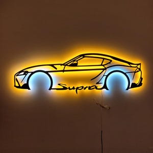 以下が含まれることがあります： 黒い金属製の壁掛けアートで、"Supra"の文字が入った車のシルエットを描いています。車の輪郭は黄色と青色のLEDライトで照らされ、光る効果を生み出しています。茶色の壁に取り付けられています。