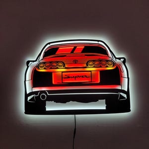May include: A black and red illuminated wall art piece showing the rear of a Supra sports car. The car's taillights and the word "Supra" are lit in red. The artwork is outlined with a white glow.