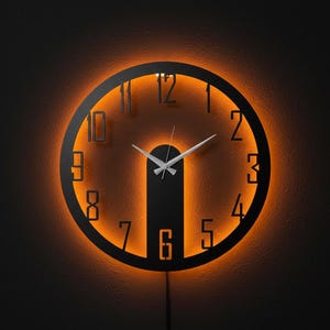 May include: A modern, round wall clock with a black frame and illuminated orange numbers. The clock hands are silver, and the numbers are cut out, allowing the orange light to shine through. The clock is mounted on a dark wall.