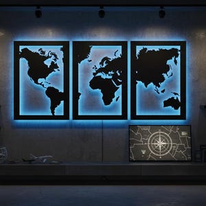 May include: A triptych of black world map wall art with blue LED backlighting. The artwork is displayed on a gray wall, with framed vintage travel posters and decorative items. A modern and stylish home decor piece.