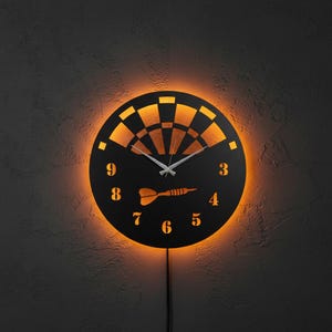 May include: A black, round wall clock with a dartboard design. The clock features a glowing orange light around the perimeter and behind the dartboard. The numbers and a dart are cut out of the black face. The clock hands are silver.