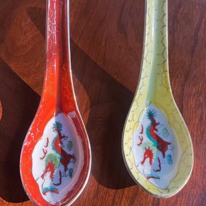 May include: Two ceramic soup spoons with long handles, one featuring a vibrant red exterior and the other a pale yellow exterior. Both spoons have a white interior bowl decorated with a colorful dragon illustration and a gold-toned rim.