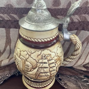 May include: A ceramic beer stein with a pewter lid and a rope-like handle. The stein features a detailed illustration of a ship with the text "Bark T. Stauon" on the bottom.