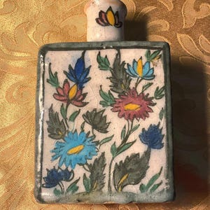May include: A rectangular ceramic bottle with a floral design. The bottle features a white background with blue, pink, and yellow flowers and green leaves. The bottle has a small, cylindrical stopper with a floral design.