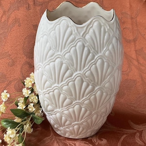 May include: A white ceramic vase with a scalloped rim and a textured, fan-like pattern. The rim has a gold trim. The vase is oval-shaped and stands on a reddish-orange surface. A sprig of yellow flowers is on the left.