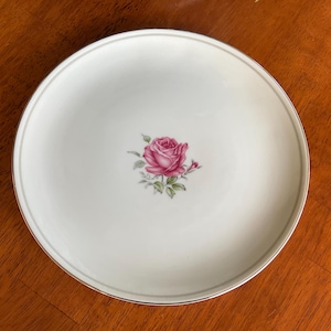May include: A white ceramic plate with a gold rim features a single pink rose with green leaves in the center.