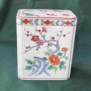 May include: A rectangular white porcelain container with a lid featuring a hand-painted floral design. The surface displays red and pink blossoms, green leaves, and blue branches. A decorative border with red and green motifs encircles the top edge.