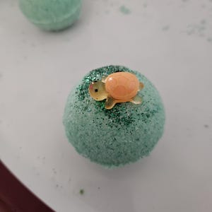 Könnte beinhalten: Eine hellgrüne Badebombe mit glitzernder Oberfläche und einer kleinen orangefarbenen Schildkrötenfigur. Die Badebombe ist rund und liegt auf einem weißen Teller. Weitere Badebomben sind im Hintergrund zu sehen.