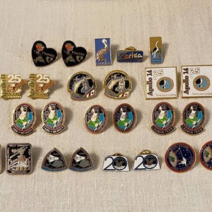May include: A collection of vintage enamel pins featuring various designs. Includes pins with the number 25, Apollo 14, Florida, and space-themed imagery. The pins are in various shapes, including hearts, squares, and circles.