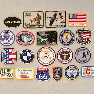 May include: A collection of embroidered patches with various designs. Patches include the American flag, a Route 66 sign, a BMW logo, and text such as "Las Vegas" and "I'll be C.B.ing you."
