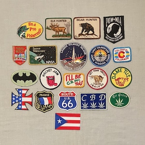 May include: A collection of twenty-one embroidered fabric patches featuring various designs including text such as I'm a Pro Fisherman, Elk Hunter, Bear Hunter, POW-MIA, Miami Beach, Space Telescope NASA, Route U.S. 66, CBD, and national flags.