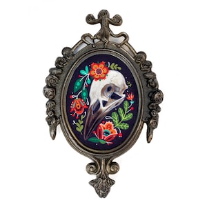 May include: A framed illustration of a bird skull with colorful flowers on a dark purple background. The frame is ornate and gold.