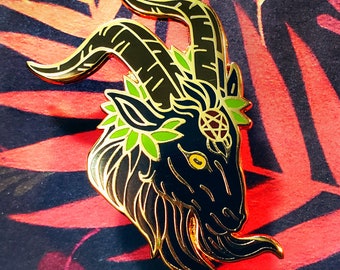 May include: A black and gold enamel pin depicting a goat's head with a green wreath and a pentagram on its forehead.