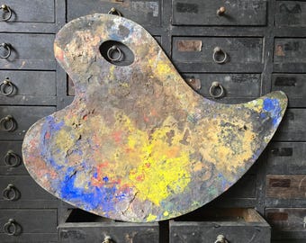 May include: A wooden artist palette with an irregular, curved shape and a thumb hole. The surface is heavily textured with dried, layered oil paint in vibrant shades of yellow, blue, red, and brown, showing significant signs of wear and use.