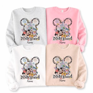 May include: Four sweatshirts in light gray, pink, white, and beige, each featuring a Disneyland graphic. The graphic includes Mickey and Minnie Mouse, Donald Duck, and Goofy, with the Disneyland logo and space for a name.