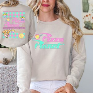 May include: Beige sweatshirt with the text "Pizza Planet" in pink and teal. The design includes a planet graphic and the words "Friday Night Kids Night" with cartoon aliens and a pizza slice.