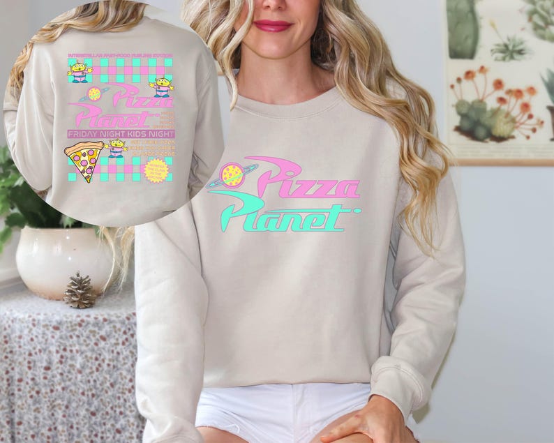 May include: Beige sweatshirt with the text "Pizza Planet" in pink and teal. The design includes a planet graphic and the words "Friday Night Kids Night" with cartoon aliens and a pizza slice.