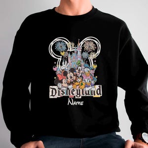 May include: Black sweatshirt featuring a colorful Disneyland graphic with Mickey Mouse, Minnie Mouse, Donald Duck, and other characters. The design includes fireworks, a castle, and the text "Disneyland" and "Name".