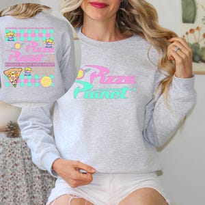 May include: A light gray sweatshirt with the words "Pizza Planet" in pink and teal. The sweatshirt has a graphic of a pizza slice, aliens, and the text "Friday Night Kids Night". The design is reminiscent of a retro cartoon.