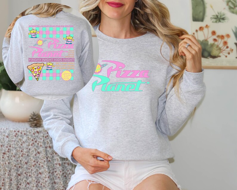 May include: A light gray sweatshirt with the words "Pizza Planet" in pink and teal. The sweatshirt has a graphic of a pizza slice, aliens, and the text "Friday Night Kids Night". The design is reminiscent of a retro cartoon.