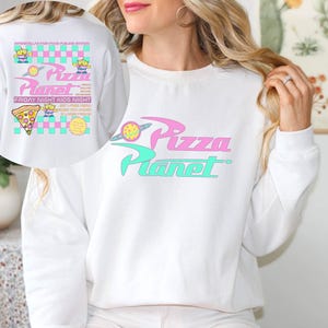 May include: White sweatshirt featuring the Pizza Planet logo in pink and teal. The design includes a planet graphic and the text "Pizza Planet". The back of the sweatshirt has additional graphics and text, including a pizza slice and alien characters.