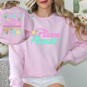May include: A light pink sweatshirt with the words "Pizza Planet" in a stylized font. The design includes a pizza slice and alien characters. The back of the sweatshirt features a similar design with the text "Friday Night Kids Night".