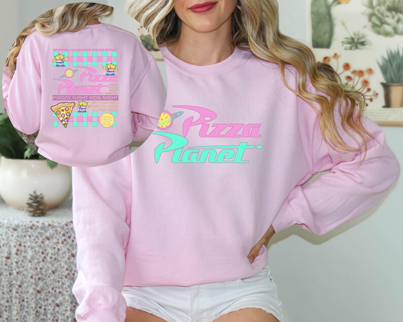 May include: A light pink sweatshirt with the words "Pizza Planet" in a stylized font. The design includes a pizza slice and alien characters. The back of the sweatshirt features a similar design with the text "Friday Night Kids Night".