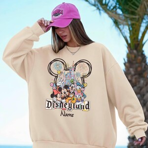 May include: Beige sweatshirt featuring a colorful Disneyland graphic with Mickey Mouse, Minnie Mouse, Donald Duck, and Goofy. The design includes a castle, fireworks, and the text "Disneyland" and "Name". The person is wearing a pink baseball cap.