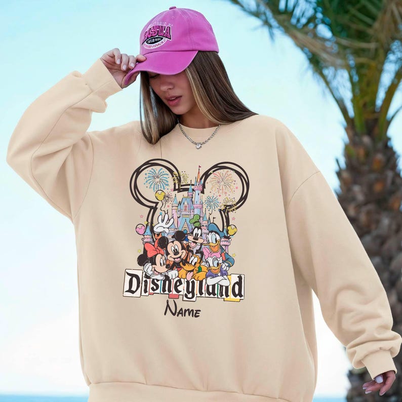 May include: Beige sweatshirt featuring a colorful Disneyland graphic with Mickey Mouse, Minnie Mouse, Donald Duck, and Goofy. The design includes a castle, fireworks, and the text "Disneyland" and "Name". The person is wearing a pink baseball cap.