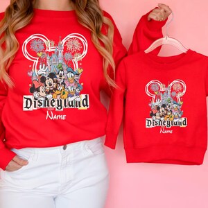 May include: Red Disneyland sweatshirts with a graphic featuring Mickey Mouse, Minnie Mouse, Donald Duck, and other Disney characters. The sweatshirts have the Disneyland logo and the word "Name" below it. One sweatshirt is adult-sized, the other is child-sized.