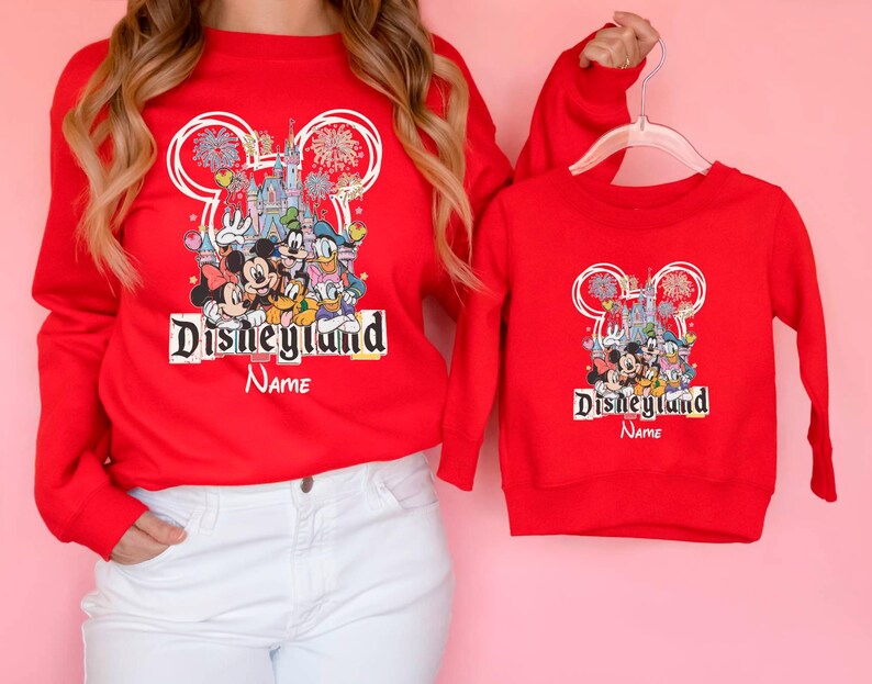 May include: Red Disneyland sweatshirts with a graphic featuring Mickey Mouse, Minnie Mouse, Donald Duck, and other Disney characters. The sweatshirts have the Disneyland logo and the word "Name" below it. One sweatshirt is adult-sized, the other is child-sized.