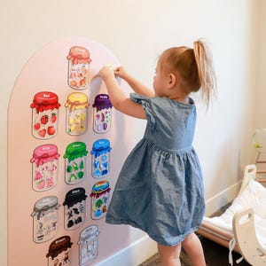 May include: A wall-mounted educational color sorting board with an arched top, featuring eleven illustrated jars labeled with color names including Orange, Red, Yellow, Purple, Pink, Green, Blue, Gray, Black, White, and Brown, each containing small matching items.