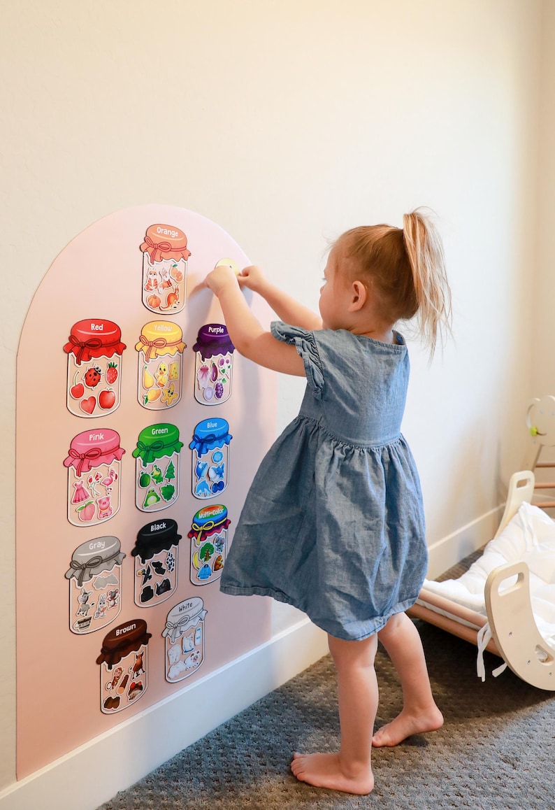 May include: A wall-mounted educational color sorting board with an arched top, featuring eleven illustrated jars labeled with color names including Orange, Red, Yellow, Purple, Pink, Green, Blue, Gray, Black, White, and Brown, each containing small matching items.