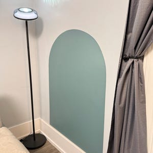 May include: A large, sage green arch-shaped wall decal applied to a white wall. The decal features a smooth, matte finish and a rounded top edge. It is positioned next to a tall, thin black floor lamp with a circular base and a white light fixture.