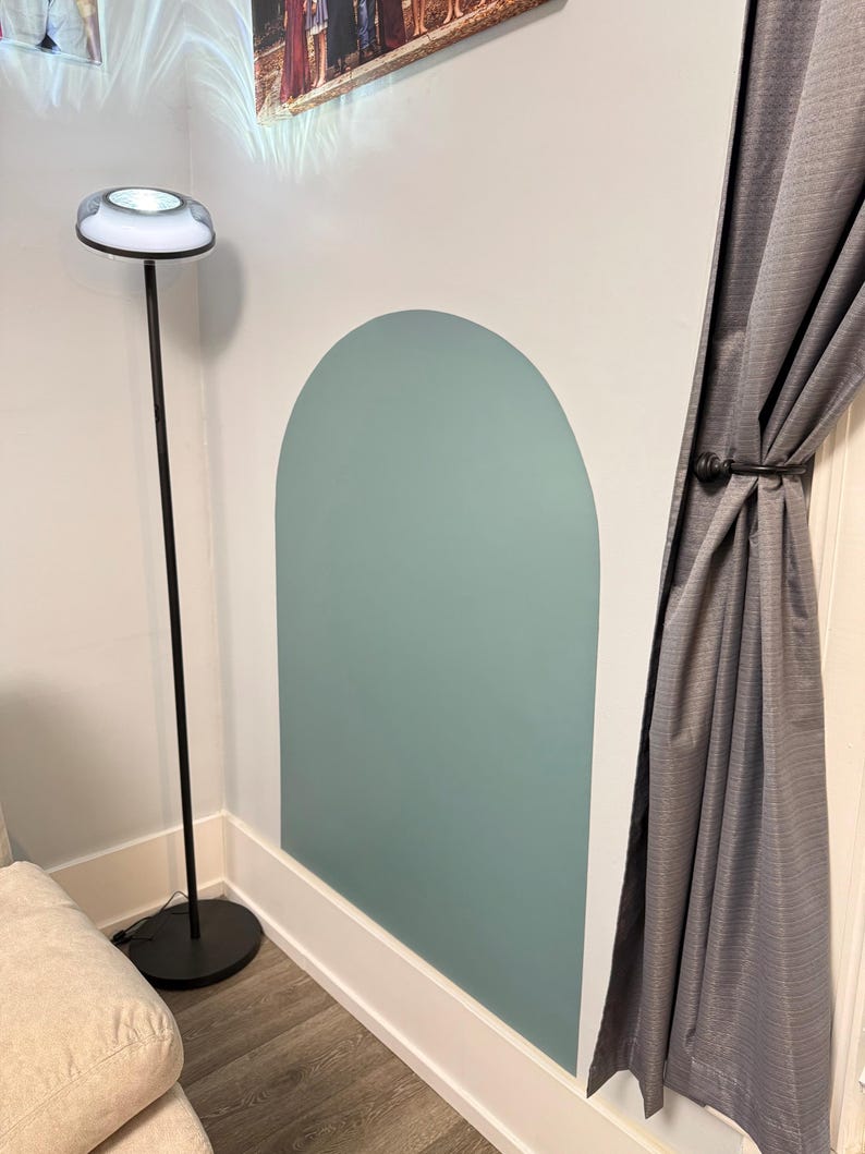 May include: A large, sage green arch-shaped wall decal applied to a white wall. The decal features a smooth, matte finish and a rounded top edge. It is positioned next to a tall, thin black floor lamp with a circular base and a white light fixture.