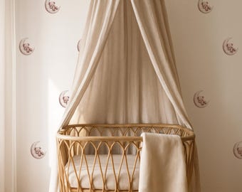 May include: A light brown wicker baby crib with a canopy and a soft blanket. The crib is set against a wall decorated with moon and teddy bear decals. The canopy and blanket are a matching beige color.
