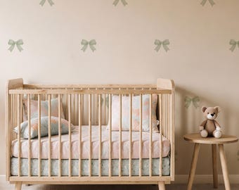 May include: A wooden crib with a pink and blue floral patterned bedding set. A small wooden side table holds a teddy bear. The wall is decorated with green bow decals. The room has a soft, neutral color palette.