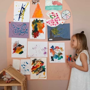 May include: A large, arched, peach-colored wall display board covered with various children's drawings, paintings, and photographs. The artwork includes colorful abstract paintings, a butterfly drawing, and a paper plate craft.