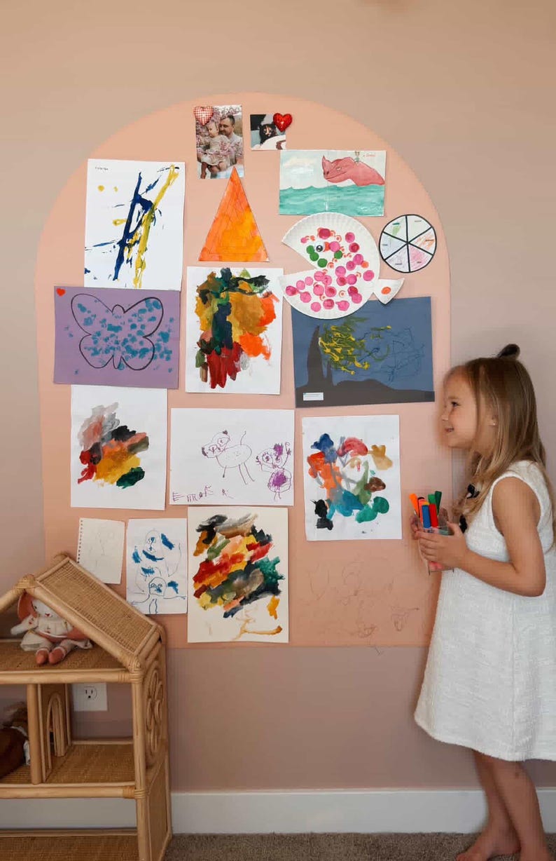 May include: A large, arched, peach-colored wall display board covered with various children's drawings, paintings, and photographs. The artwork includes colorful abstract paintings, a butterfly drawing, and a paper plate craft.