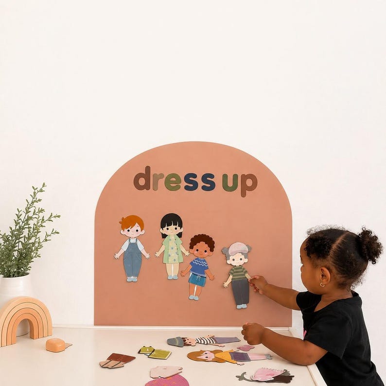 May include: A terracotta-colored arched wall decal with the text "dress up" in dark letters. Four illustrated paper dolls are attached to the surface, while various paper clothing items are scattered on the white surface below the arch.