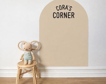 May include: A beige arch-shaped wall decal featuring the black text CORA'S CORNER, positioned above a light-colored wooden chair with a woven seat and a small plush bear wearing light green overalls sitting on the chair seat.