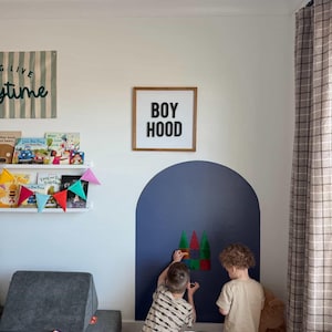 May include: A square wooden sign with a white background and black text reading BOY HOOD, mounted on a wall above a dark blue painted arch. The sign features a simple frame and is displayed in a room with a bookshelf and children playing nearby.