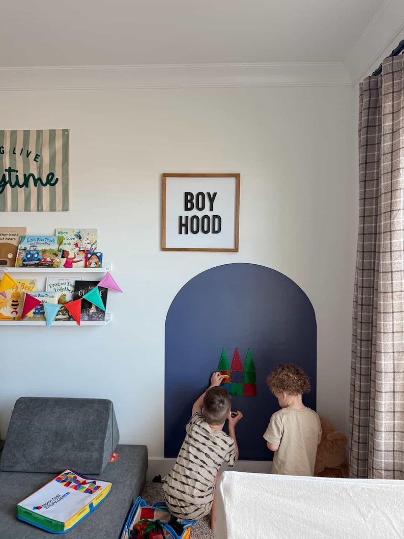 May include: A square wooden sign with a white background and black text reading BOY HOOD, mounted on a wall above a dark blue painted arch. The sign features a simple frame and is displayed in a room with a bookshelf and children playing nearby.