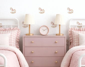 May include: A child's bedroom with two white metal beds, each with pink bedding and ruffled gingham pillows. A pink dresser with a clock and lamps sits between the beds. Rocking horse decals adorn the white wall, creating a soft, inviting space.