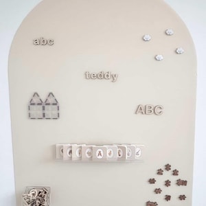 May include: A beige arched display board featuring wooden letters spelling abc, teddy, and ABC, four small cloud shapes, a house-shaped magnetic tile structure, a row of small illustrated booklets on a shelf, a clear bin of letters, and puzzle pieces.