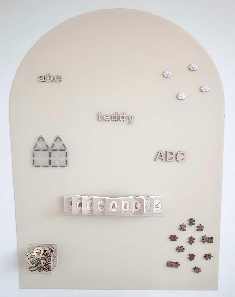 May include: A beige arched display board featuring wooden letters spelling abc, teddy, and ABC, four small cloud shapes, a house-shaped magnetic tile structure, a row of small illustrated booklets on a shelf, a clear bin of letters, and puzzle pieces.