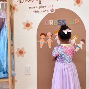 May include: A wall decal set featuring a brown arch, the text DRESS UP, and various paper doll figures. The wall also displays the text kind of chick to... make playtime this cute, surrounded by orange and brown flower decals on a white wall.